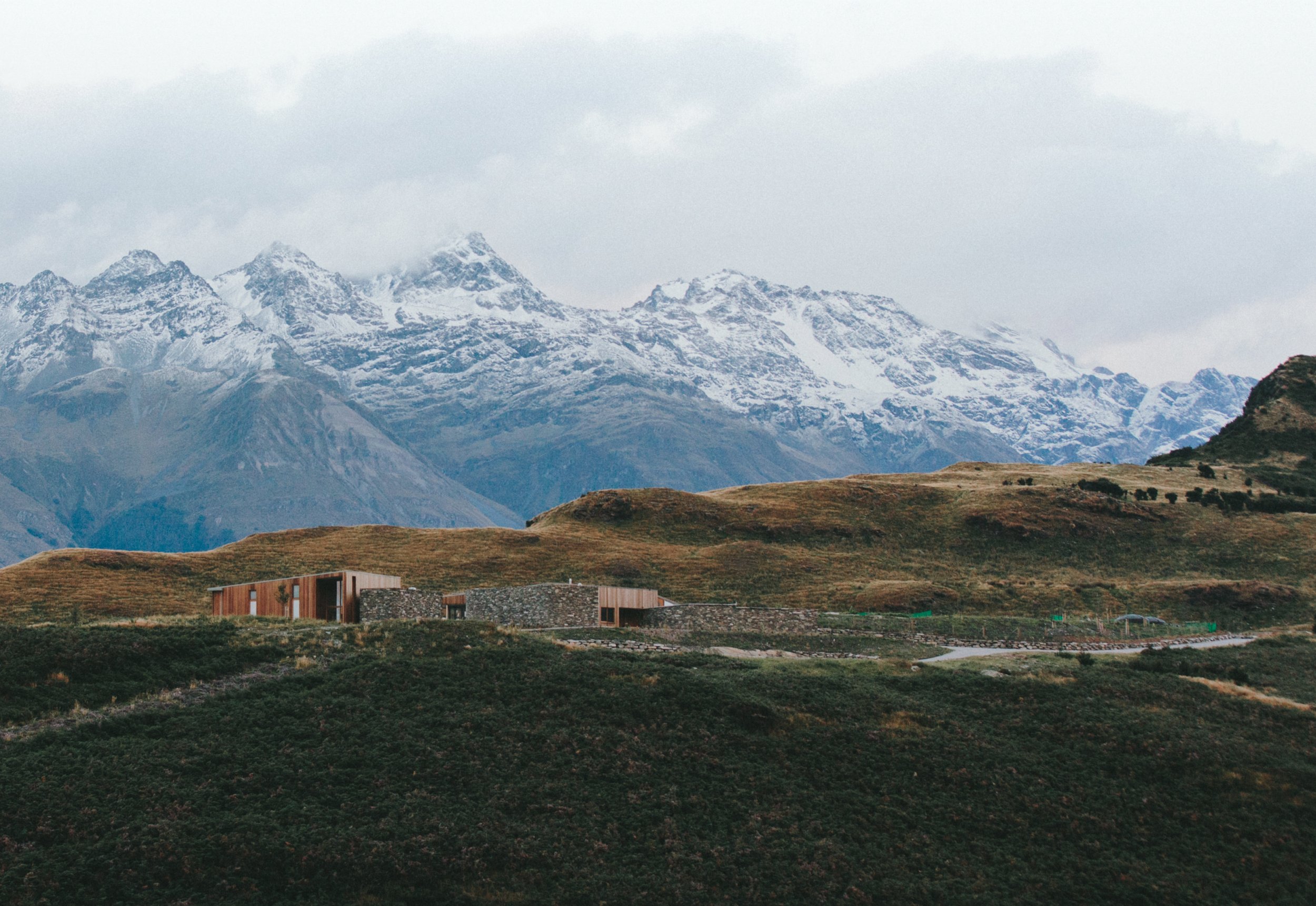 Modern house with wooden and stone exterior walls located on a hillside with grassy terrain, snow-capped mountains in the background under a cloudy sky.